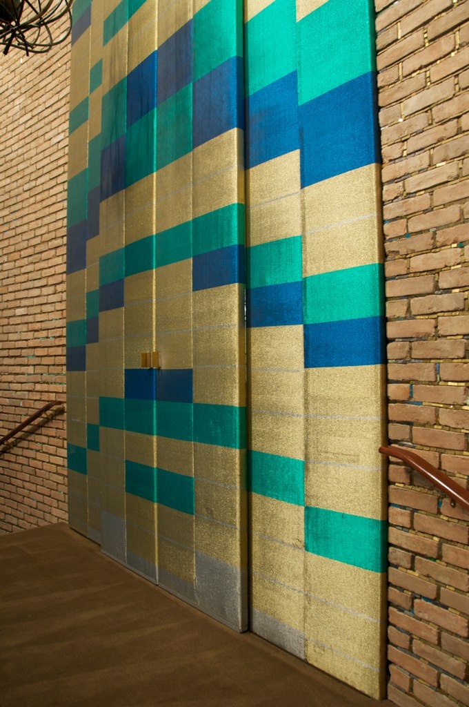 Tall textile panels with a geometric pattern of gold, teal and blue blocks set between brick walls