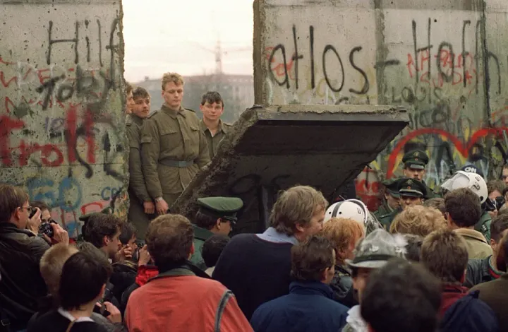 East German border guards looking through a gap in the Berlin Wall as crowds gather, 1989